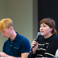 three college student panelists sitting at the front of a conference room, one speaking while the others listen